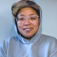 Light brown round Filipine/x person wearing glasses, a blue sweater, floral hat, and headphones.